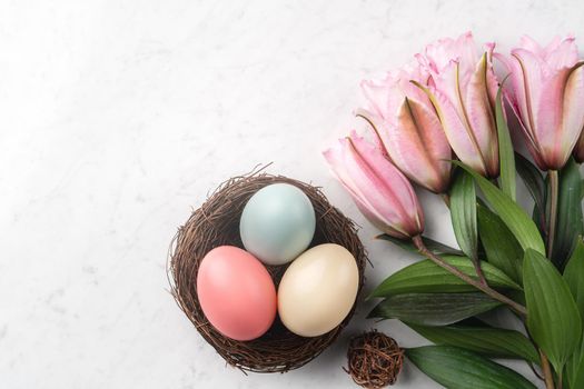 Colorful Easter eggs in the nest with pink lily flower on bright marble white table background.