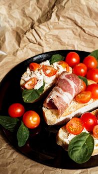 Three bruschetta on a black plate on craft paper. Appetizer with cherry tomatoes, curd cheese, spinach and bacon. Vertical photo