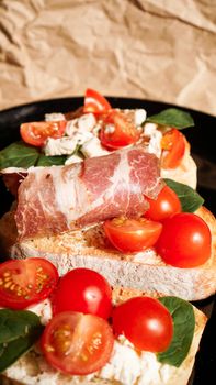 Three bruschetta on a black plate on craft paper. Appetizer with cherry tomatoes, curd cheese, spinach and bacon. Vertical photo