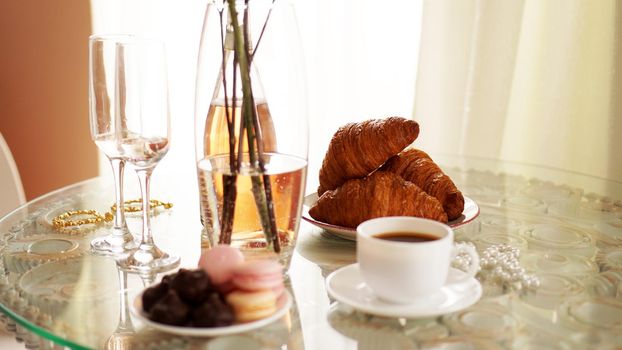 Glass table with a cup of coffee, sweet croissants. On the table is a bottle of wine and glasses. A festive and romantic photo. Morning after da