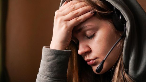 Portrait of a young girl in a hoodie and with a headset. Her eyes are closed and her hand is on her forehead. Overwork. Tired girl streamer or call center worker.