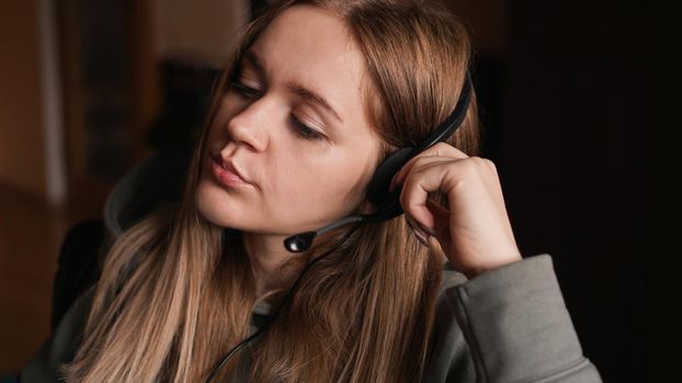 Portrait of a young girl in a hoodie and with a headset. She listens to the client with a tired look. Monotonous work. Call center worker. Remote work from home. Brown tones pictures