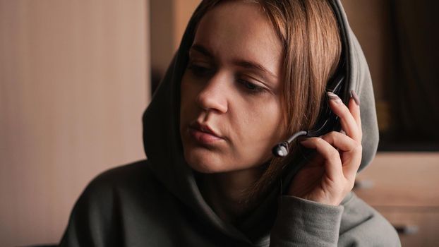 Portrait of a young girl in a hoodie and with a headset. Call center worker. She holds the earpiece to hear the client better. Communication problems. Remote work from home. Brown tones pictures