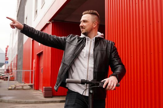 Young attractive man on electric scooter over red background. A man showing a direction with his hand