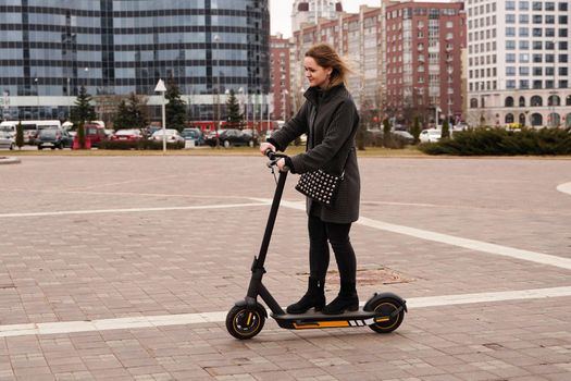 Beautiful girl in a coat rides electric scooter around the city on a sunny day