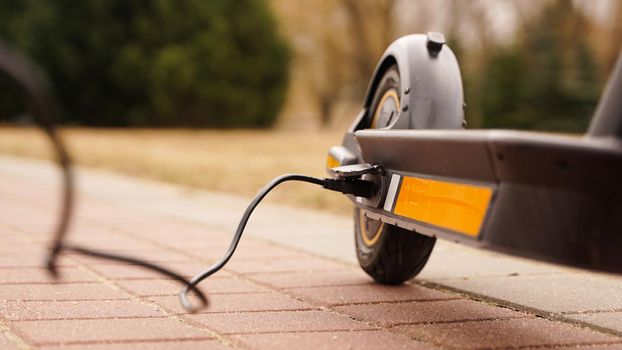 The electric scooter is charging outdoors. The wire runs into the camera. Ecological transport