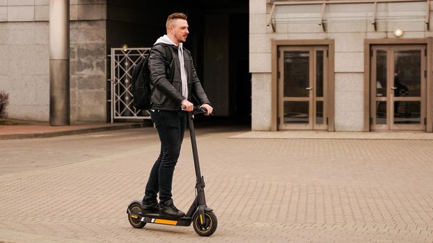 Tall man on an electric scooter against the backdrop of gray city buildings. Guy in a black leather jacket