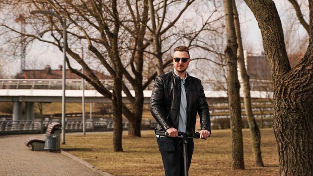 Young man in a black jacket and sunglasses rides an electronic scooter in a spring park on a sunny day