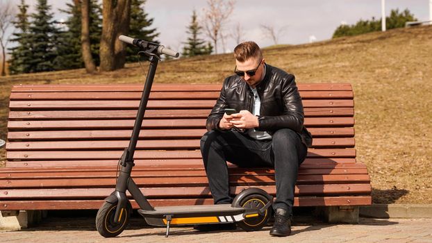 Young attractive man sits on a bench on a spring day. Electric scooter next to a bench. Man holds a smartphone in his hands.
