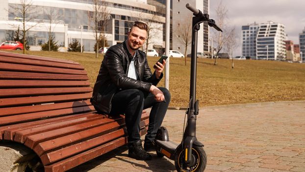 Young attractive man sits on a bench on a spring day. Electric scooter next to a bench. Man holds a smartphone in his hands. He looks at the camera and smiles