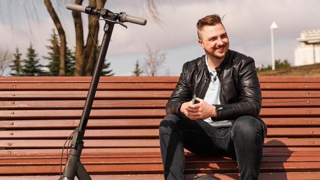 Young attractive man sits on a bench on a spring day. Electric scooter next to a bench. Man holds a smartphone in his hands. He looks away and smiles