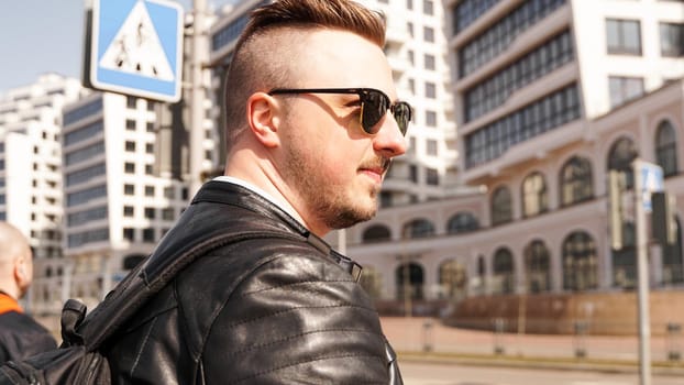 An attractive man wearing black leather jacket with dark sunglasses against the backdrop of a glass building in the city
