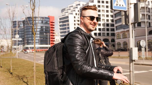 A smiling guy in sunglasses stands at the crossroads waiting for the green light of a traffic light. The steering wheel of an electric scooter in his hands.