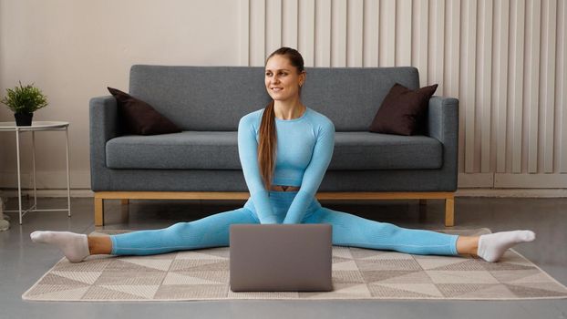 A beautiful woman in a blue tracksuit is stretching at home in front of a laptop. She sits in a transverse twine.