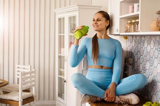 A woman sits on the countertop in the kitchen and drinks water from a sports bottle. Healthy lifestyle concept.