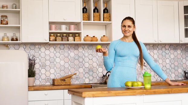 A beautiful woman in a blue suit is standing in the kitchen, smiling and holding a green apple. The concept of sports and weight loss at home.