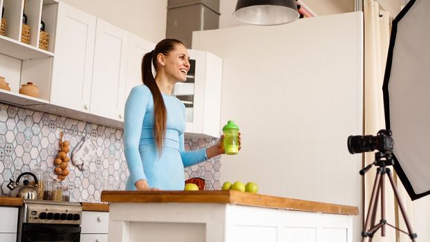 Healthy woman recording her video blog about healthy food additives while standing at kitchen. She is holding a bottle of sports nutrition and smiling