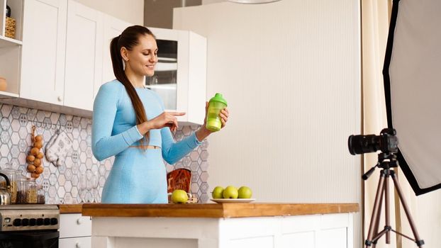 Healthy woman recording her video blog about healthy food additives while standing at kitchen. She is holding a bottle of sports nutrition and smiling