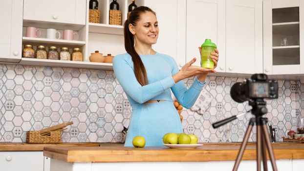 Healthy woman recording her video blog about healthy food additives while standing at kitchen. She is holding a bottle of sports nutrition and smiling