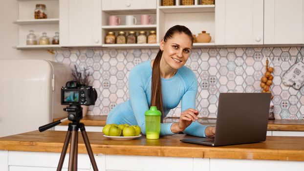 Young woman in kitchen with laptop smiling. Food blogger concept. A woman is recording a video about healthy eating. Camera on a tripod.