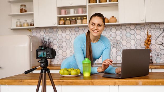 Young woman in kitchen with laptop smiling. Food blogger concept. A woman is recording a video about healthy eating. Camera on a tripod.