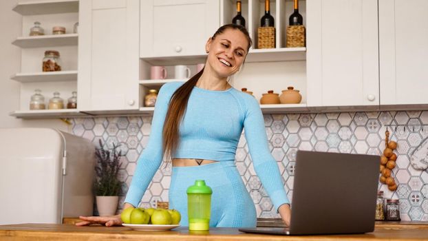 Smiling woman using computer in modern kitchen interior. Cooking and healthy lifestyle concept. Woman broadcasts online and gestures emotionally