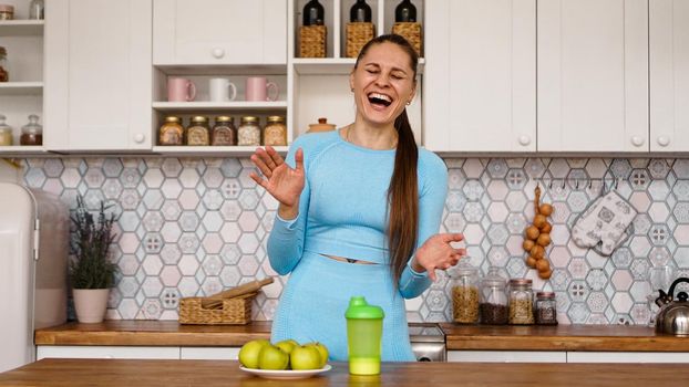 Athletic woman talks about healthy eating in the kitchen and laughs. On the table are green apples and a green bottle for a sports drink and water