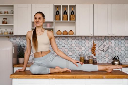 Athletic woman in sportswear posing on the table top of a light modern kitchen. The concept of beauty, health, proper nutrition.