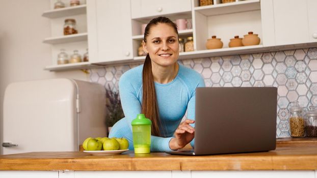 Smiling woman using computer in modern kitchen interior. Cooking and healthy lifestyle concept. A woman is looking at the camera and smiling