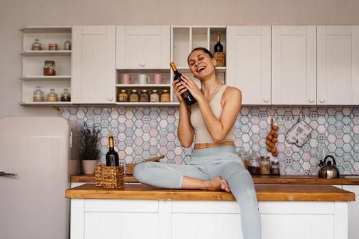 Athletic woman in a tracksuit in a light kitchen drinks red wine from a bottle after doing sports. Woman laughs
