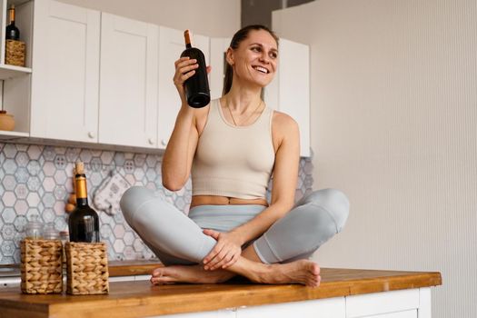 Athletic woman in a tracksuit in a light kitchen drinks red wine from a bottle after doing sports.