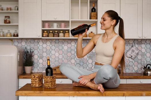 Athletic woman in a tracksuit in a light kitchen drinks red wine from a bottle after doing sports.