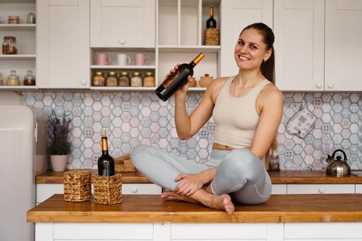 Athletic woman in a tracksuit in a light kitchen drinks red wine from a bottle after doing sports. Woman smiling and looking at the camera