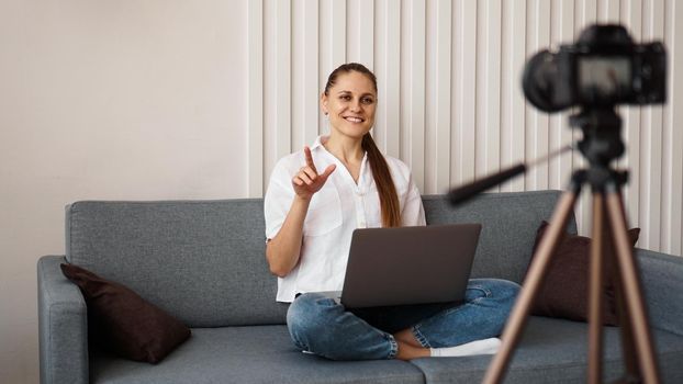 Smiling female blogger records a new video. She sits on the couch at home and holds a laptop. Positive business blog concept.