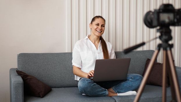 Smiling female blogger records a new video. She sits on the couch at home and holds a laptop. Positive business blog concept.