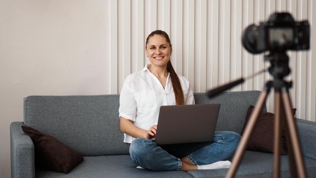 Smiling female blogger records a new video. She sits on the couch at home and holds a laptop. Positive business blog concept.