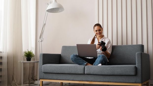 Happy young woman with photo camera using laptop at home. The photographer looks at the pictures taken and sits on the couch.