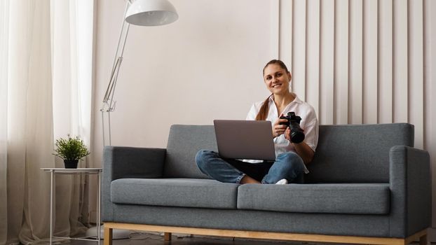 Happy young woman with photo camera using laptop at home. The photographer is holding the camera and smiling.