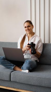Happy young woman with photo camera using laptop at home. The photographer is holding the camera and smiling. Vertical photo.