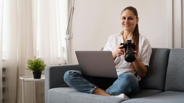 Happy young woman with photo camera using laptop at home. The photographer is holding the camera and smiling.