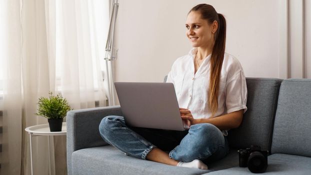Happy young woman using laptop at home. On the couch next to the woman is a camera. Photographer retouches photographs at home.