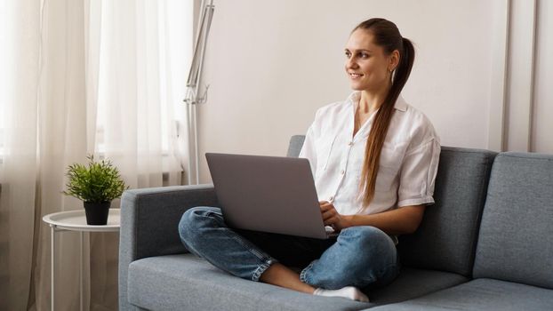 Smiling happy woman sitting on the sofa and using laptop. The concept of remote work or study during quarantine.