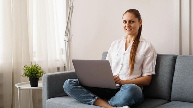 Smiling happy woman sitting on the sofa and using laptop. The concept of remote work or study during quarantine.