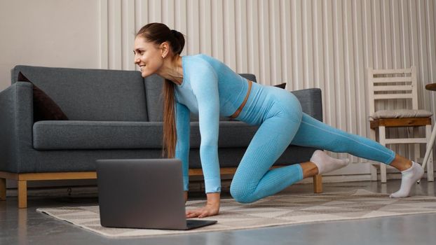 Beautiful young woman in a blue suit doing fitness exercise at home. She looks at her laptop and repeats the movements for the online course.