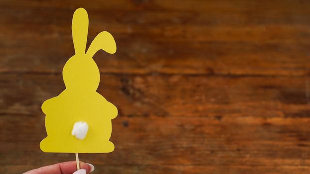 Yellow rabbit made of paper on a toothbrush. Hand holds Easter baking decor. Easter card on wooden background