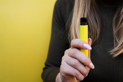 Yellow disposable electronic cigarette in a female hand. Bright yellow background. Melon, pineapple or lemon flavored vape