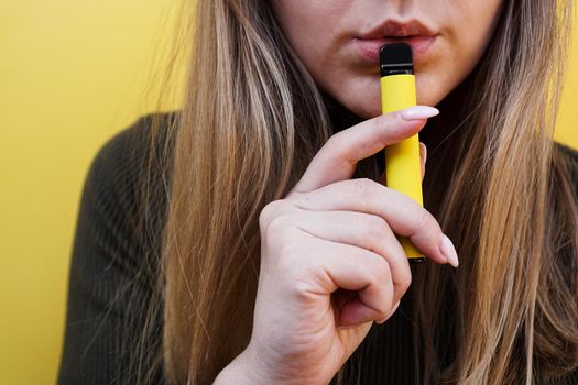 Close up of a young girl smokes a disposable electronic cigarette. Bright yellow background. Alternative to regular cigarettes and vape