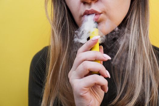 Close up of a young girl smokes a disposable electronic cigarette and exhales smoke. Bright yellow background. Alternative to regular cigarettes and vape