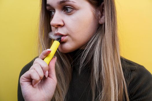 Close up of a young girl smokes a disposable electronic cigarette. Bright yellow background. Alternative to regular cigarettes and vape