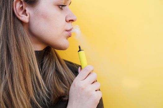 Close up of a young girl smokes a disposable electronic cigarette and exhales smoke. Bright yellow background. Alternative to regular cigarettes and vape